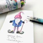 watercolor pencil painting of a holiday gnome with white beard wearing a red cap and watercolor pencils, waterbrush, and pen around drawing