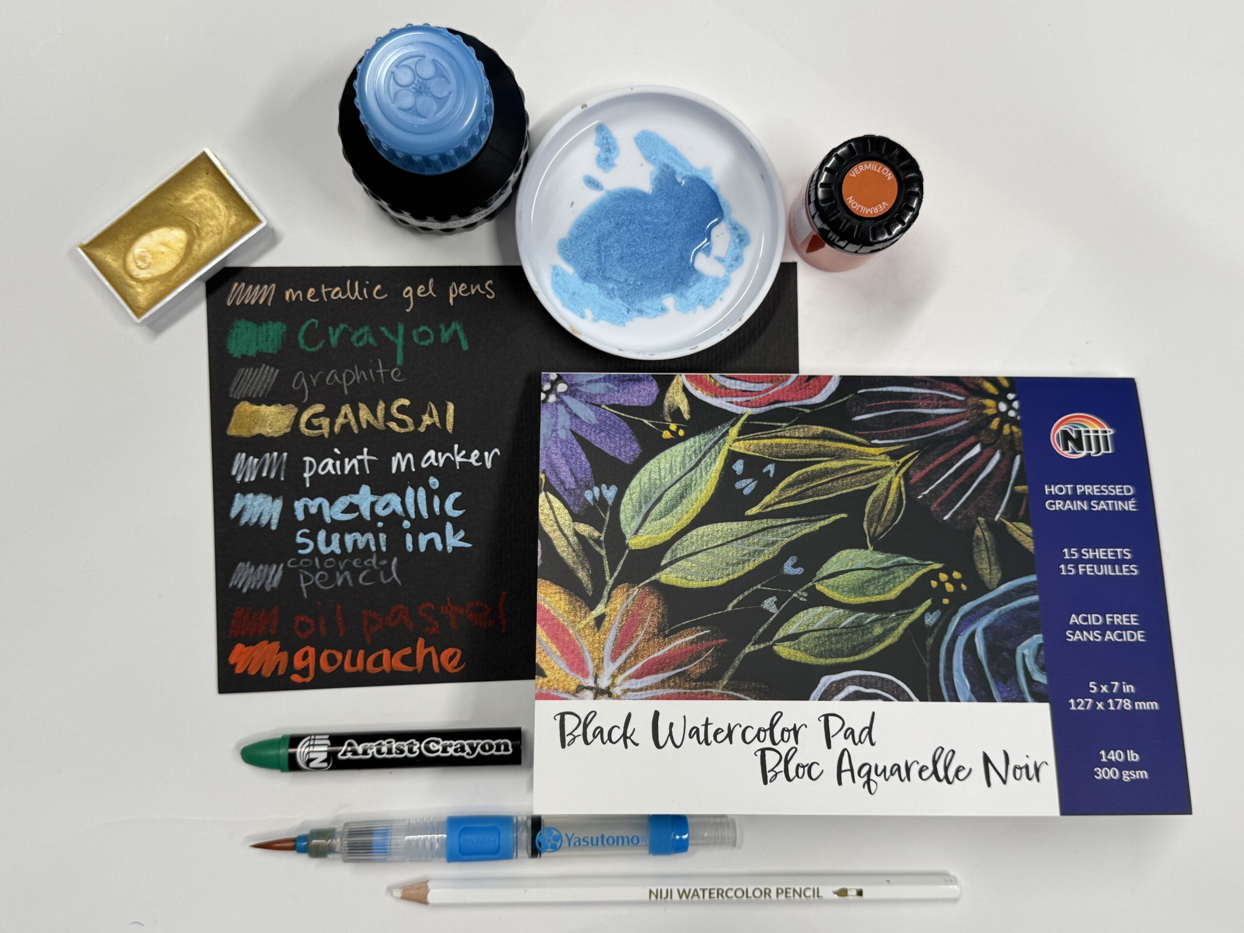a flat lay spread of black watercolor paper with swatches, black watercolor paper pad, metallic inks, crayon, pencil, brush, and other art supplies