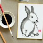 shikishi board with a black and white painting of a rabbit, with paint brushes, and ink dish, and a colorful watercolor painting surrounding it
