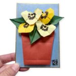 shikishi board with blue background and origami pansies in a pot, held in a hand