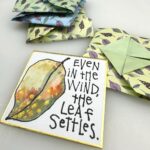 small square shikishi board with a gansai leaf and quote "Even in the wind, the leaf settles." and origami tato envelopes in the background