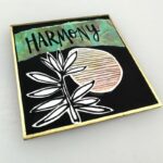 square shikishi board colored black with paper cutouts of an abstract botanical shape. a circle and the word Harmony across the top