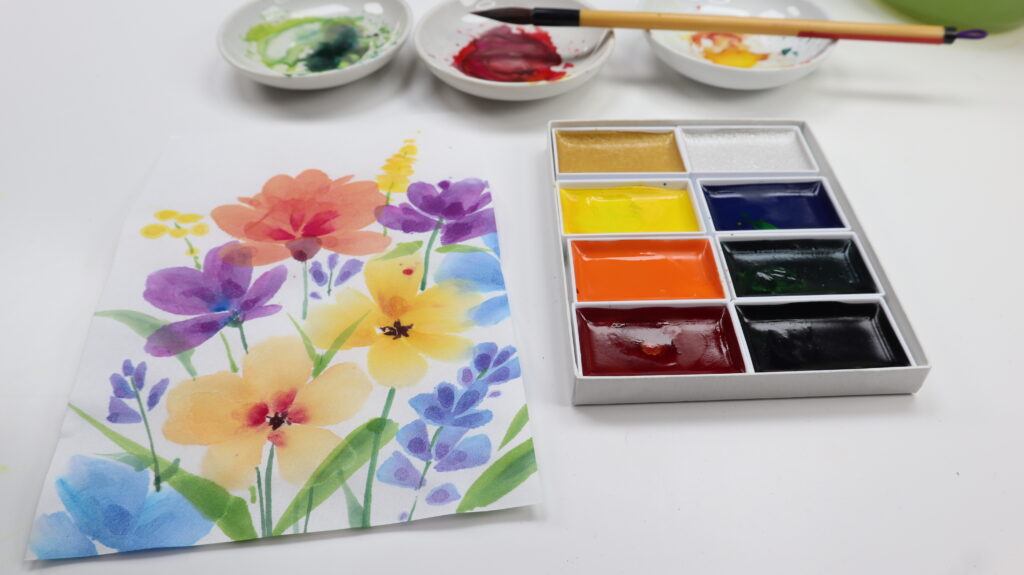 colorful gansai flowers painting next to a gansai set and three porcelain dishes with paint in them and a brush laid over the saucers