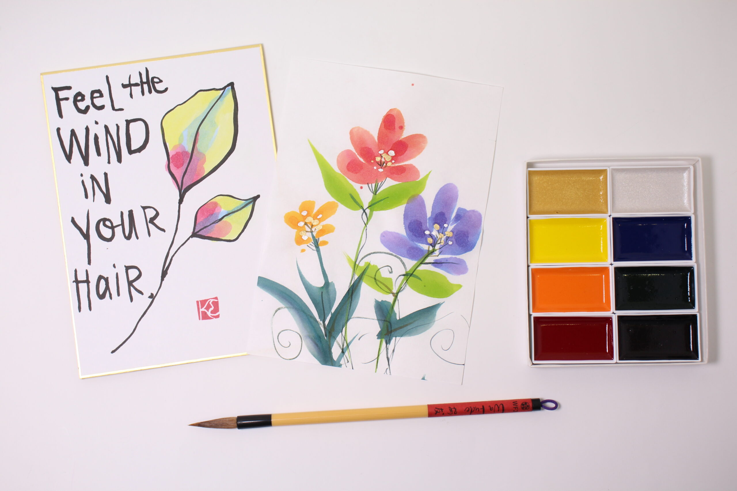 leaf painting on a shikishi board with the words "Feel the wind in your hair" and a floral gansai painting on Hosho paper next to 8 gansai colors in a box and a Wa Fude brush