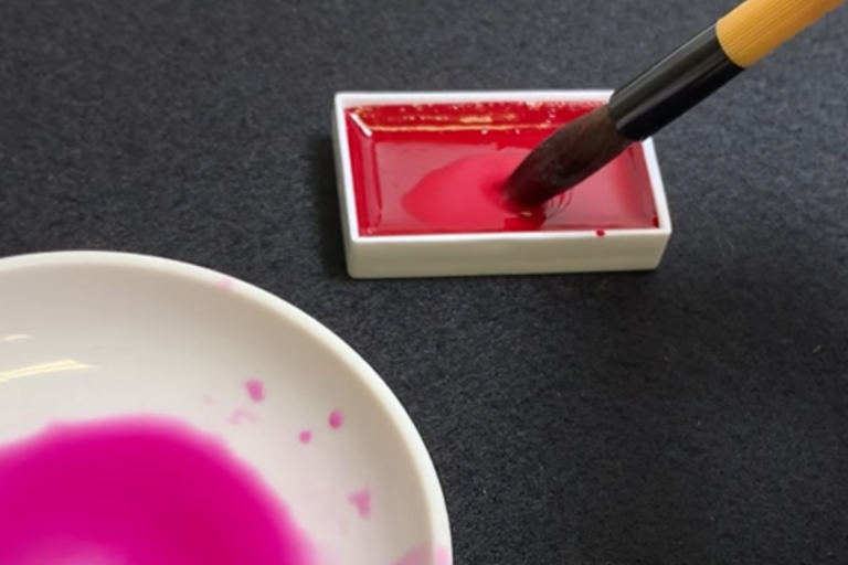 brush being dipped directly into red gansai paint with a porcelain dish with thinned color to the side