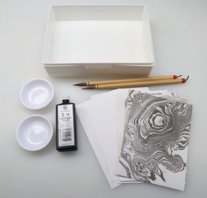 rectangular white plastic tray, two round dishes, bottle of sumi ink, two sumi brushes, and a spread of Hosho paper with a suminagashi print against a white background