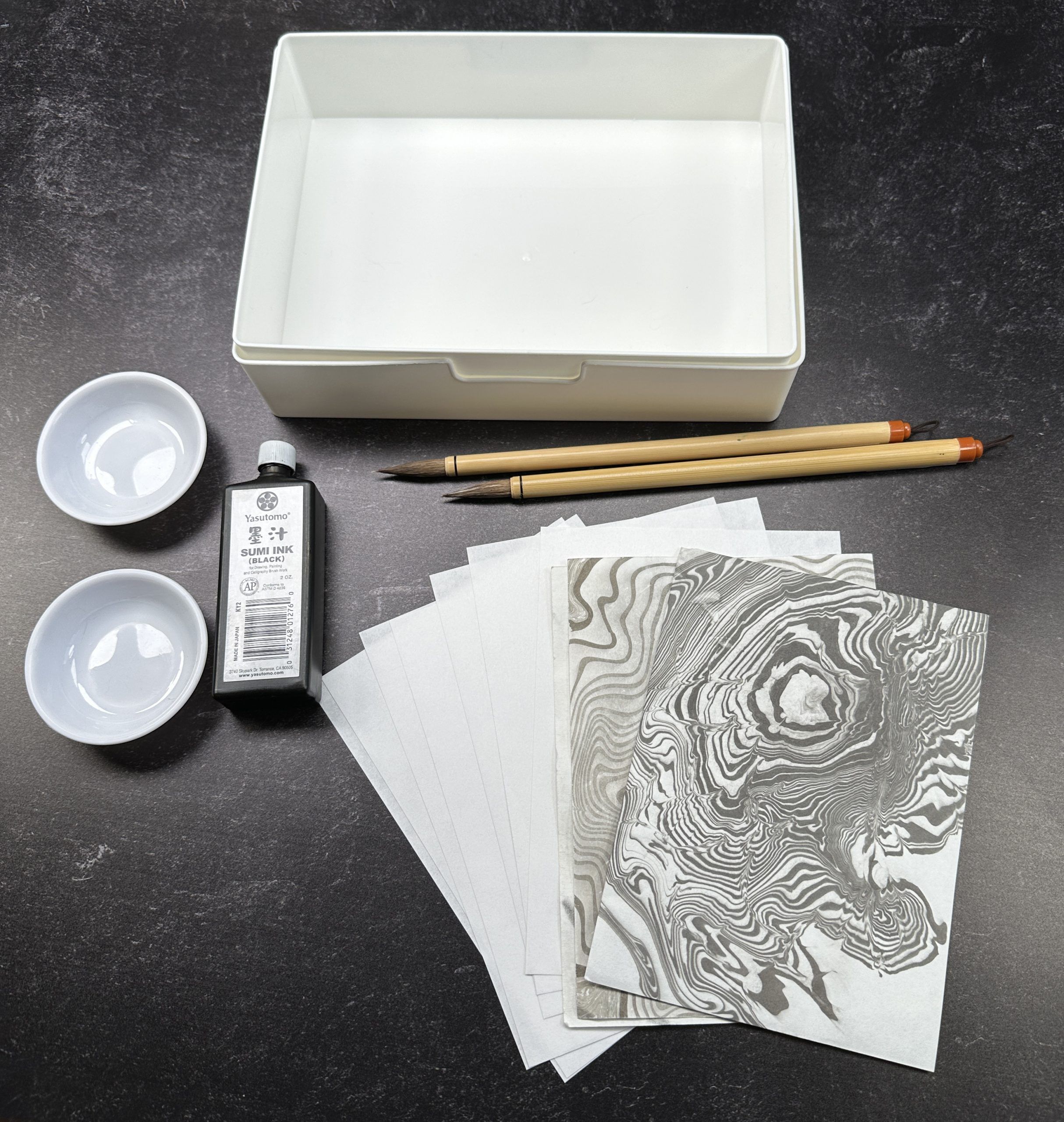 water tray, two brushes, two round dishes, small liquid black ink bottle, white washi papers and sheets of paper with suminagashi Japanese water marbling designs against a black background