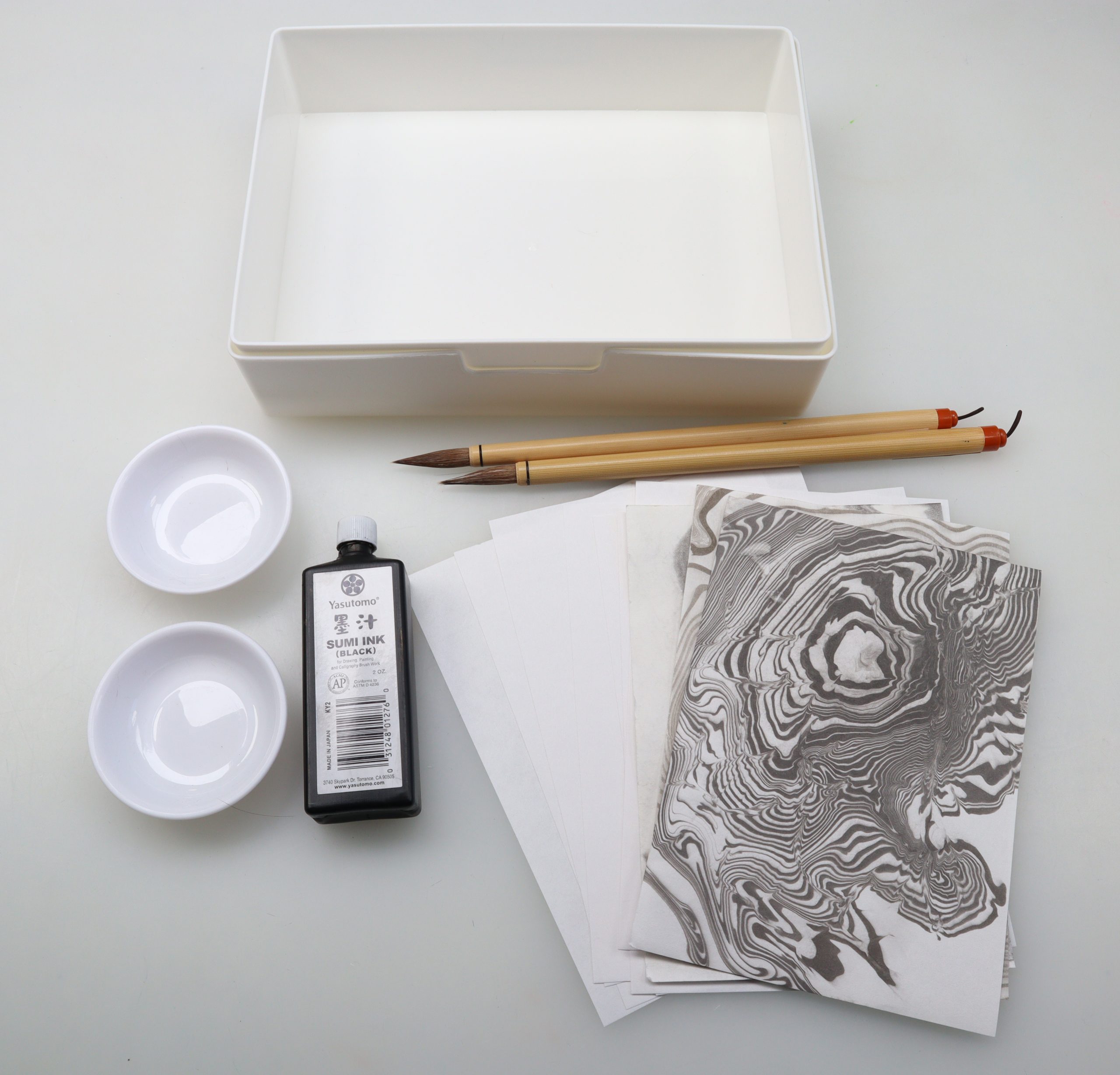 water tray, two brushes, two round dishes, small liquid black ink bottle, white washi papers and sheets of paper with suminagashi Japanese water marbling designs against a black background