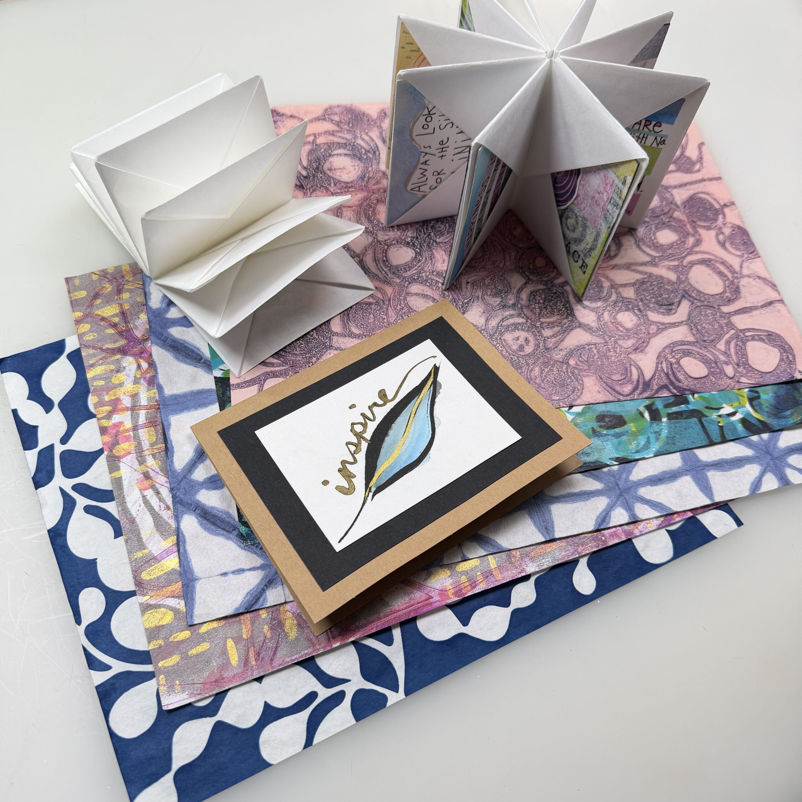 spread of colorful printed papers with a painted card and two folded books arranged on top