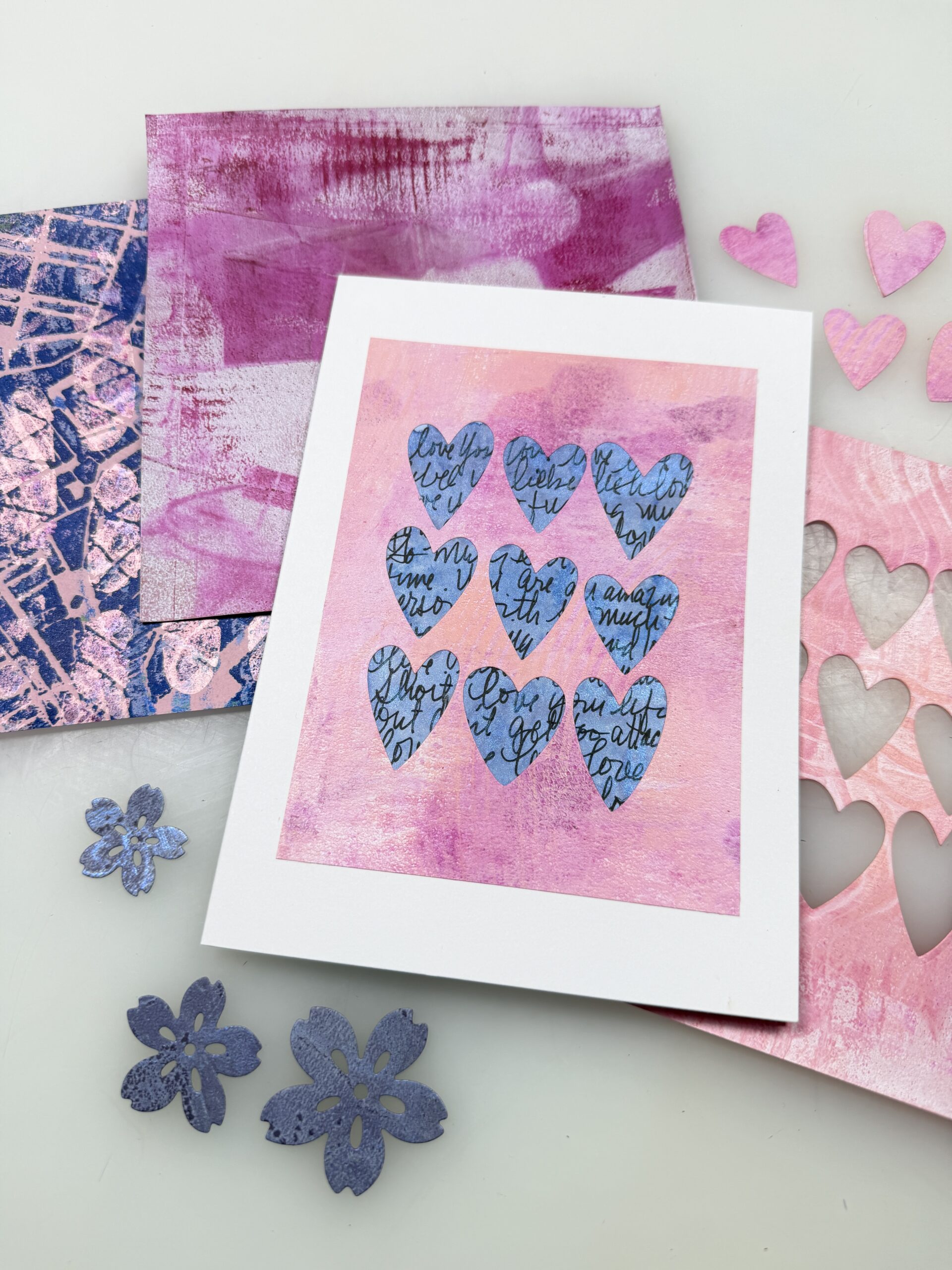 card with gel printed pink sheet with heart shaped cutouts and a blue decorative sheet in the background, surrounded by other printed papers in the background