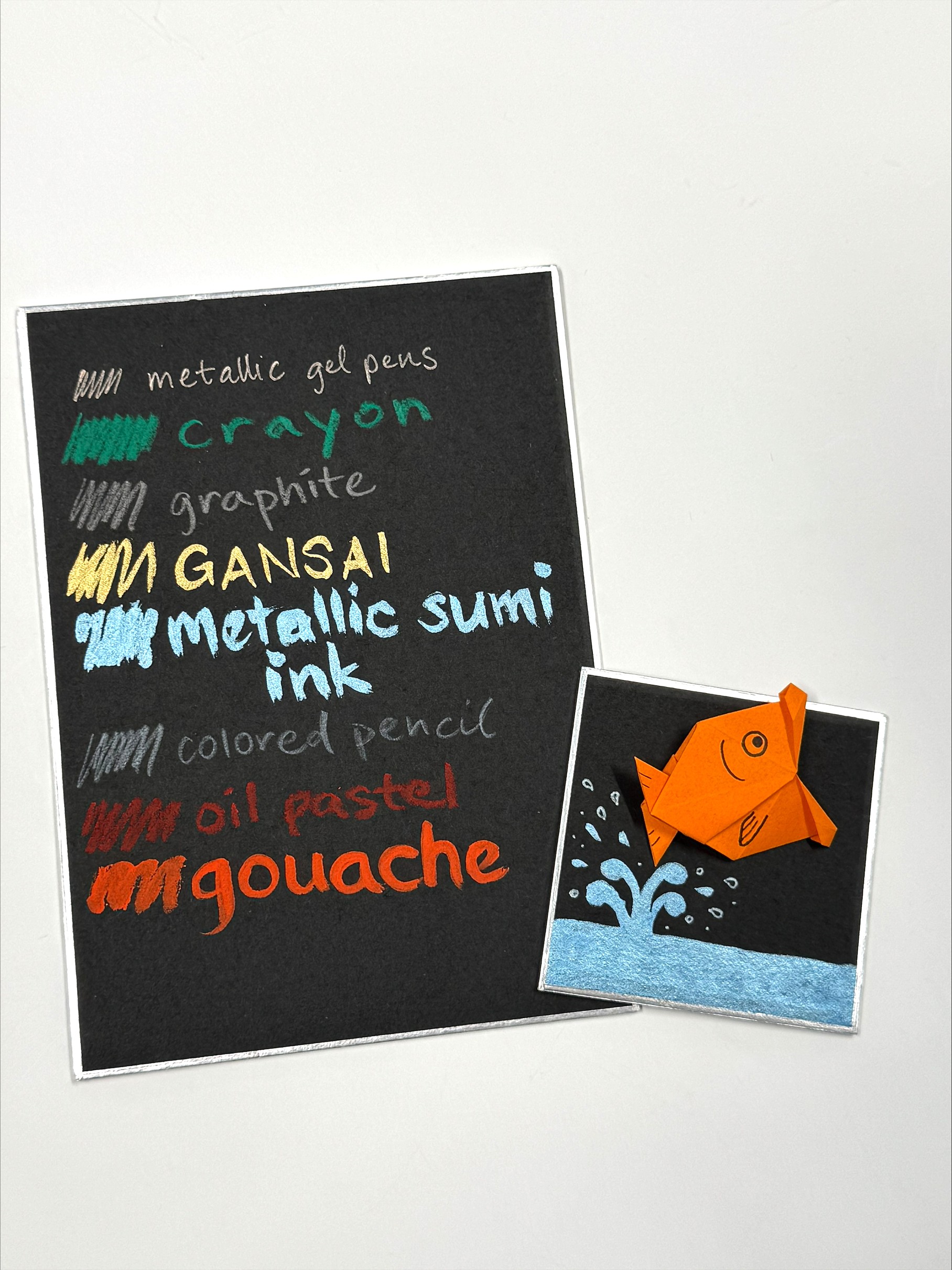 black shikishi board with swatches of different mediums and smaller black shikishi board with origami goldfish jumping out of water