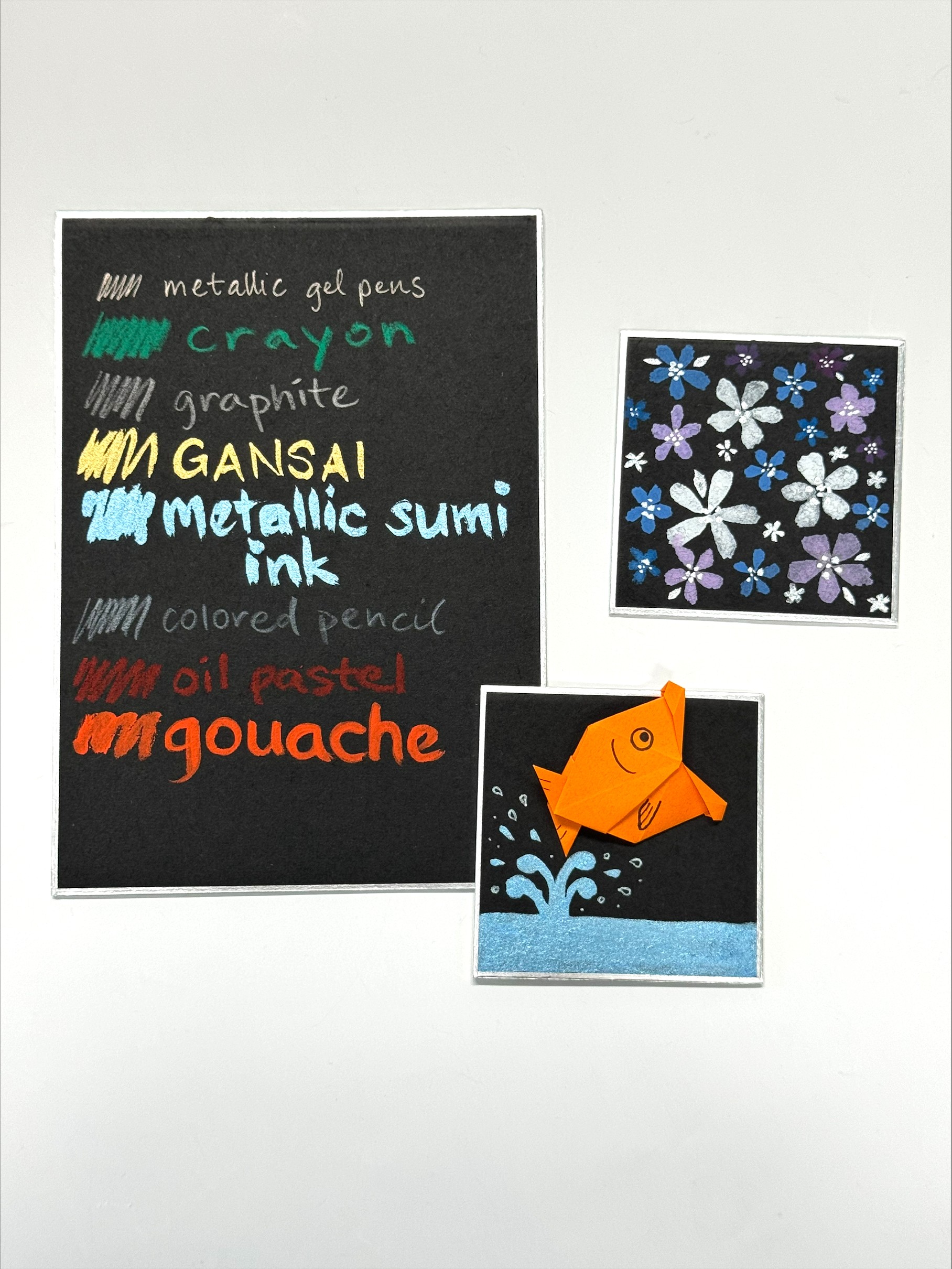 3 black shikishi boards featuring swatches of different mediums, gansai painted flowers, and an origami goldfish jumping out of water